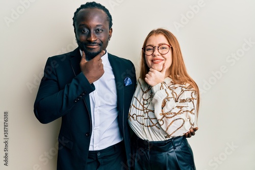 Young interracial couple wearing business and elegant clothes looking confident at the camera smiling with crossed arms and hand raised on chin. thinking positive.