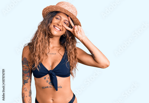 Young hispanic woman with tattoo wearing bikini and summer hat doing peace symbol with fingers over face, smiling cheerful showing victory