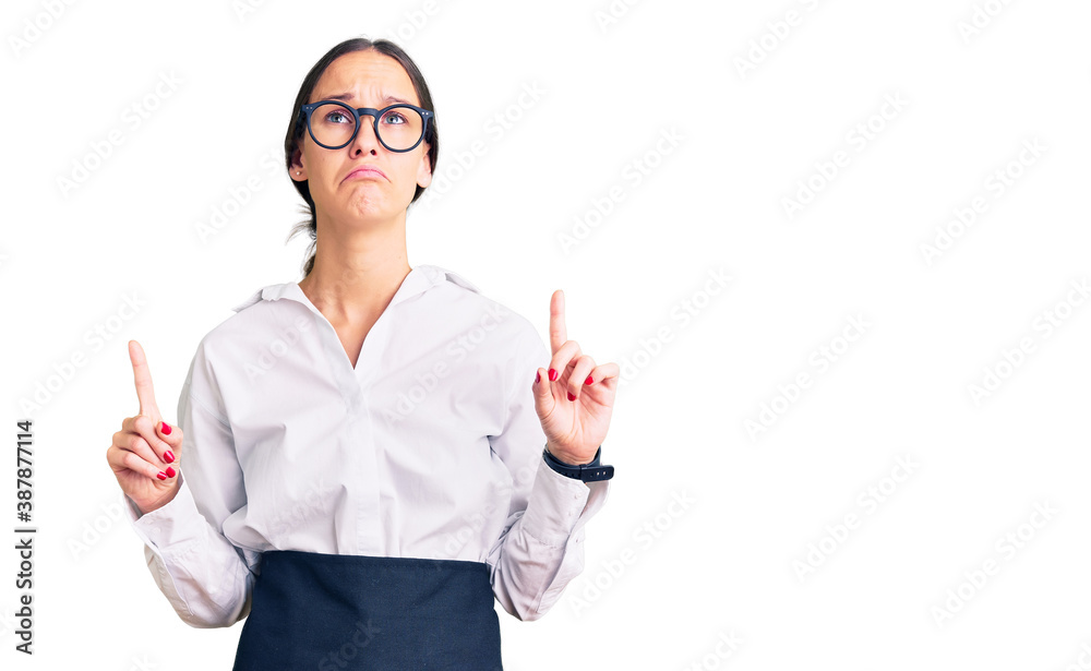 Beautiful brunette young woman wearing professional waitress apron pointing up looking sad and upset, indicating direction with fingers, unhappy and depressed. Beautiful brunette young woman wearing professional waitress apron pointing up looking sad and upset, indicating direction with fingers, unhappy and depressed.