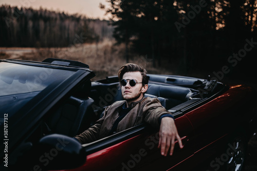  brutal man with a beard sits in a red convertible in sunglasses in nature. Male bachelor and driver style.
