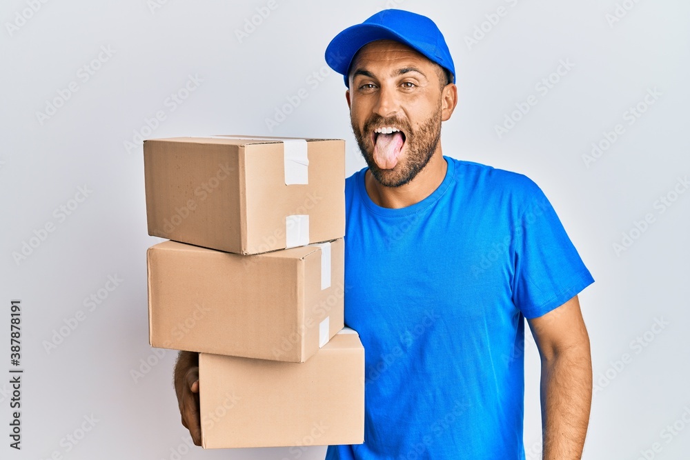 Handsome man with beard wearing courier uniform holding delivery packages sticking tongue out happy with funny expression. emotion concept. Handsome man with beard wearing courier uniform holding delivery packages sticking tongue out happy with funny expression. emotion concept.