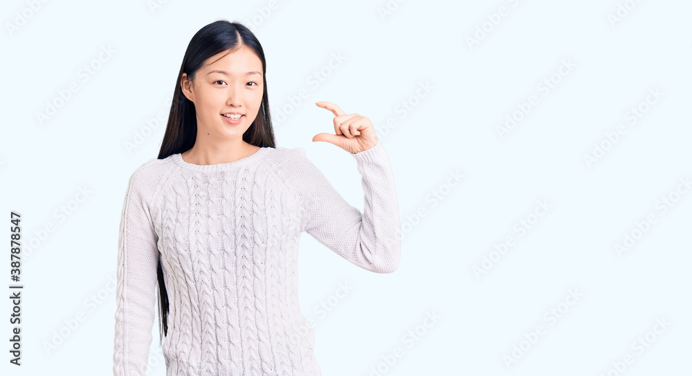 Young beautiful chinese woman wearing casual sweater smiling and confident gesturing with hand doing small size sign with fingers looking and the camera. measure concept. Young beautiful chinese woman wearing casual sweater smiling and confident gesturing with hand doing small size sign with fingers looking and the camera. measure concept.