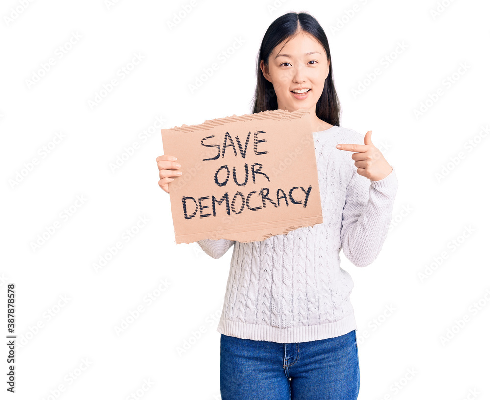 Young beautiful chinese woman holding save our democracy cardboard banner smiling happy pointing with hand and finger Young beautiful chinese woman holding save our democracy cardboard banner smiling happy pointing with hand and finger