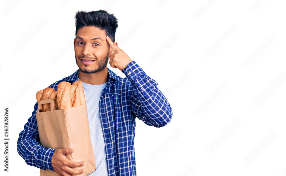 Handsome latin american young man holding paper bag with bread smiling pointing to head with one finger, great idea or thought, good memory Handsome latin american young man holding paper bag with bread smiling pointing to head with one finger, great idea or thought, good memory