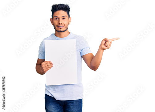 Handsome latin american young man holding cardboard banner with blank space smiling happy pointing with hand and finger to the side