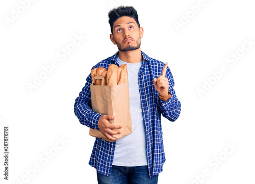 Handsome latin american young man holding paper bag with bread pointing up looking sad and upset, indicating direction with fingers, unhappy and depressed.