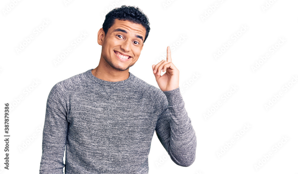 Hispanic handsome young man wearing casual sweater pointing finger up with successful idea. exited and happy. number one.
