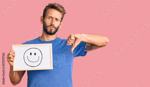 Handsome blond man with beard holding blackboard with happy face with angry face, negative sign showing dislike with thumbs down, rejection concept