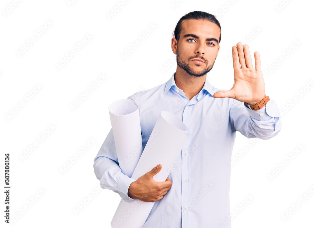 Young handsome man holding paper blueprints with open hand doing stop sign with serious and confident expression, defense gesture Young handsome man holding paper blueprints with open hand doing stop sign with serious and confident expression, defense gesture