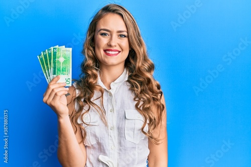 Young blonde girl holding norwegian krone banknotes looking positive and happy standing and smiling with a confident smile showing teeth photo