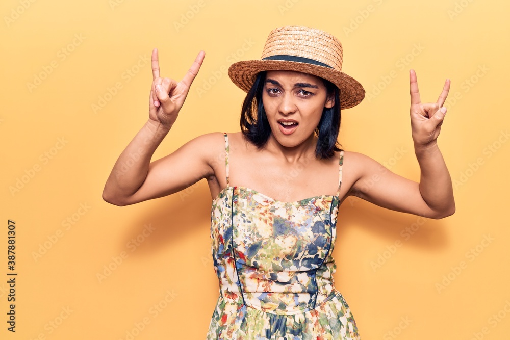 Young beautiful latin woman wearing summer hat shouting with crazy expression doing rock symbol with hands up. music star. heavy concept. Young beautiful latin woman wearing summer hat shouting with crazy expression doing rock symbol with hands up. music star. heavy concept.