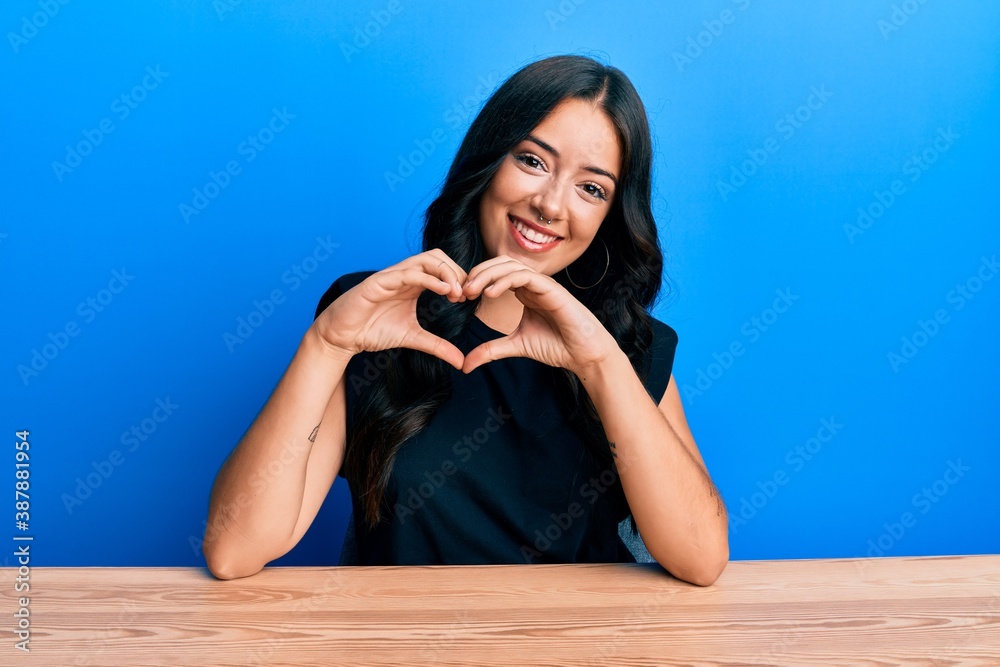 Beautiful brunette young woman wearing casual clothes sitting on the table smiling in love doing heart symbol shape with hands. romantic concept. Beautiful brunette young woman wearing casual clothes sitting on the table smiling in love doing heart symbol shape with hands. romantic concept.