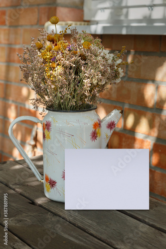 Postcard mockup with old enameled jug and dried flowers. Empty card with space for text, print or lettering. Vintage wooden table and brick wall.