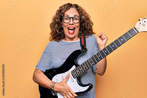 Middle age beautiful musician woman playing electric guitar over isolated yellow background screaming proud, celebrating victory and success very excited with raised arm