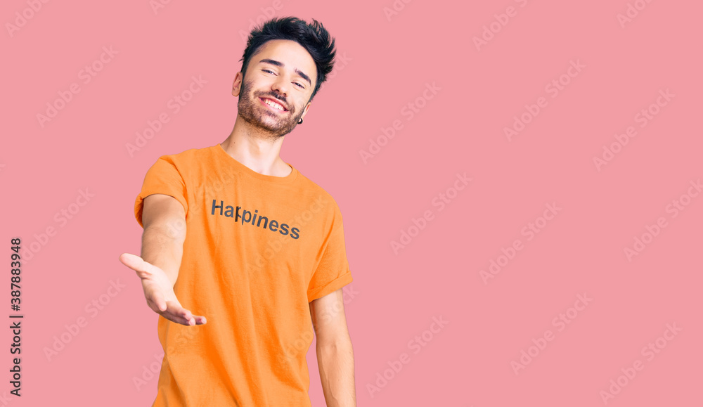 Young hispanic man wearing t shirt with happiness word message smiling cheerful offering palm hand giving assistance and acceptance.
