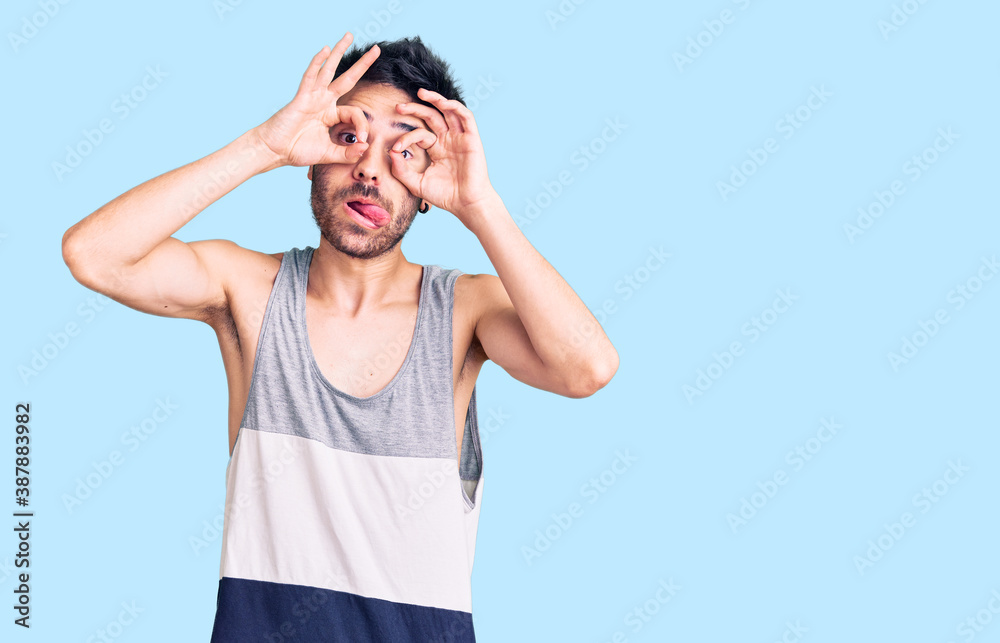 Young hispanic man wearing casual clothes doing ok gesture like binoculars sticking tongue out, eyes looking through fingers. crazy expression. Young hispanic man wearing casual clothes doing ok gesture like binoculars sticking tongue out, eyes looking through fingers. crazy expression.