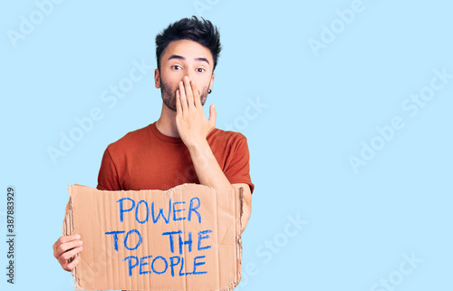 Young hispanic man holding power to the people banner covering mouth with hand, shocked and afraid for mistake. surprised expression © Krakenimages.com