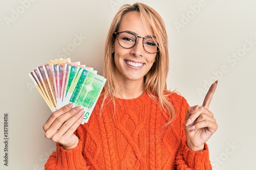 Fotografie Beautiful blonde woman holding norwegian krone banknotes smiling happy pointing
