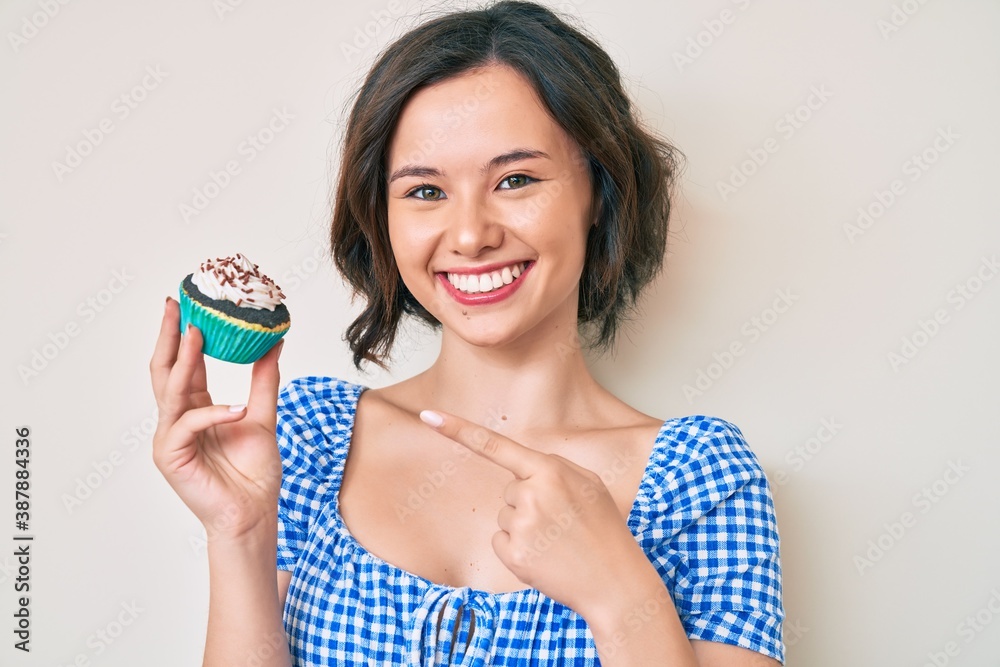 Young beautiful woman holding cupcake smiling happy pointing with hand and finger Young beautiful woman holding cupcake smiling happy pointing with hand and finger