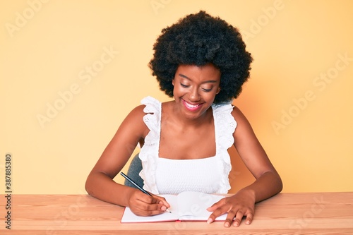 Young african american woman writing book looking positive and happy standing and smiling with a confident smile showing teeth