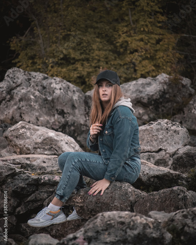 Brunette girl wearing a jeans outfit and a black cap sitting on a rock in the nature, looking at the camera in the distance with a shocked surprised look