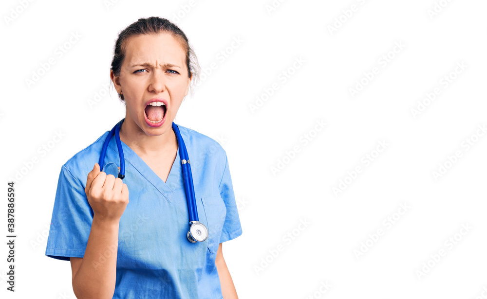 Young beautiful blonde woman wearing doctor uniform and stethoscope angry and mad raising fist frustrated and furious while shouting with anger. rage and aggressive concept. Young beautiful blonde woman wearing doctor uniform and stethoscope angry and mad raising fist frustrated and furious while shouting with anger. rage and aggressive concept.