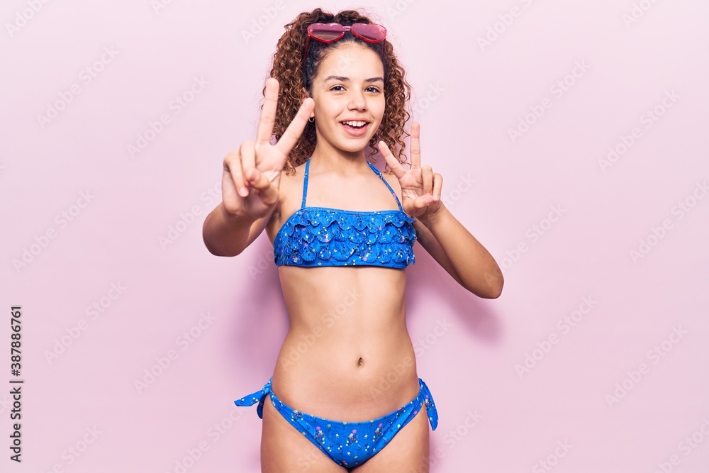 Beautiful kid girl with curly hair wearing bikini and sunglasses smiling looking to the camera showing fingers doing victory sign. number two. - obrazy, fototapety, plakaty 