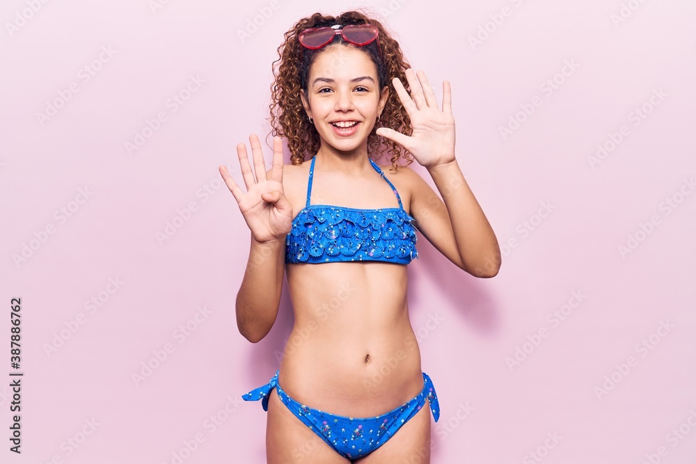 Naklejka premium Beautiful kid girl with curly hair wearing bikini and sunglasses showing and pointing up with fingers number nine while smiling confident and happy.