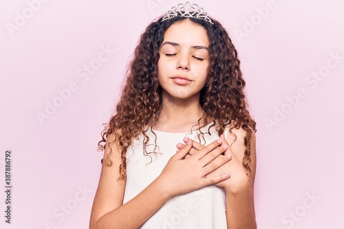 Beautiful kid girl with curly hair wearing princess tiara smiling with hands on chest, eyes closed with grateful gesture on face. health concept.