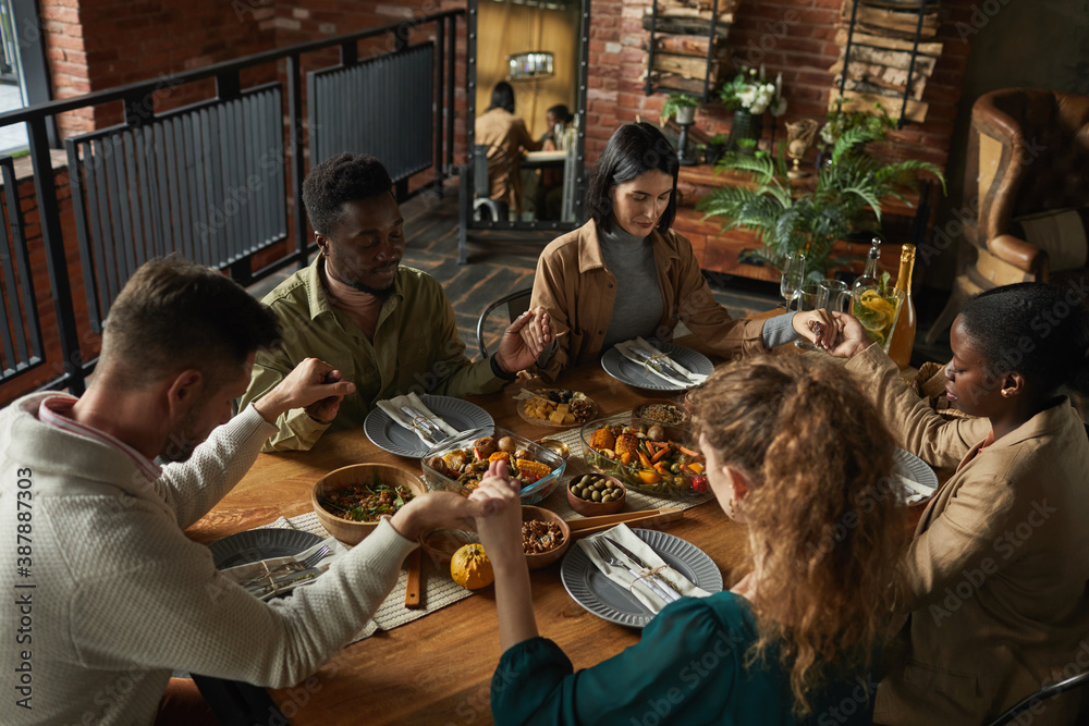 High angle portrait of multi-ethnic group of elegant young people praying and holding hands while sitting at dinner table during Thanksgiving celebration, copy space