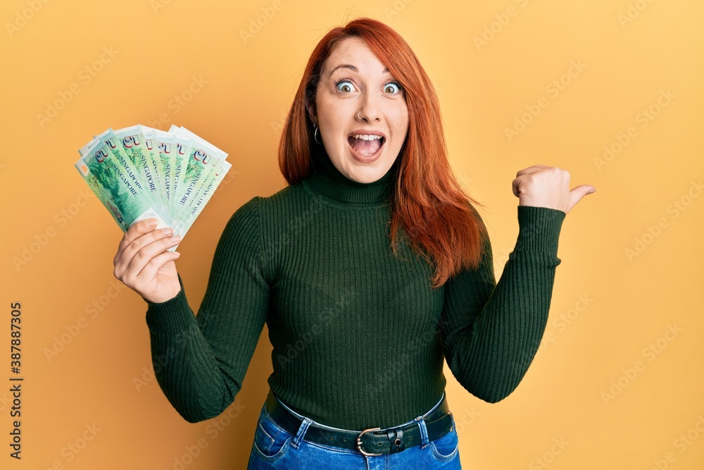 Beautiful redhead woman holding 5 singapore dollars banknotes pointing thumb up to the side smiling happy with open mouth Beautiful redhead woman holding 5 singapore dollars banknotes pointing thumb up to the side smiling happy with open mouth