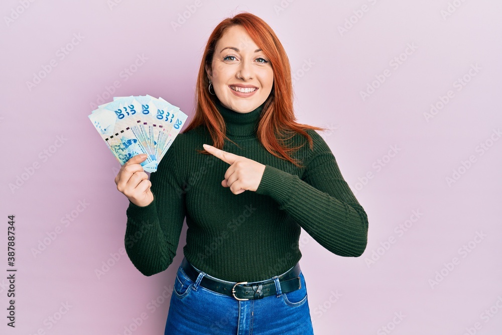 Beautiful redhead woman holding 20 hong kong dollars banknotes smiling happy pointing with hand and finger Beautiful redhead woman holding 20 hong kong dollars banknotes smiling happy pointing with hand and finger