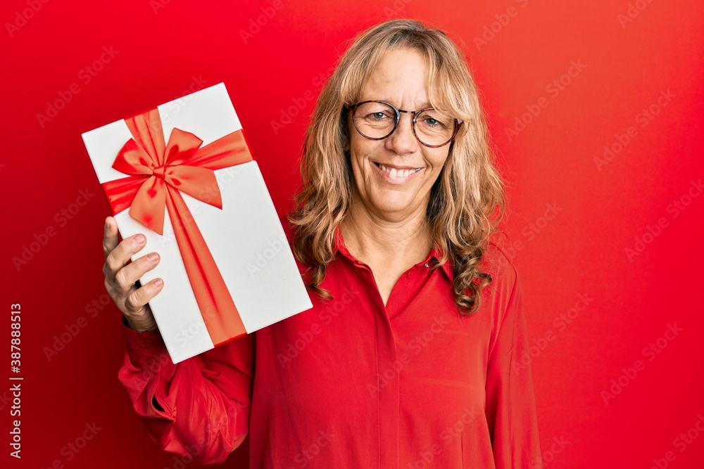 Middle age blonde woman holding gift looking positive and happy standing and smiling with a confident smile showing teeth Middle age blonde woman holding gift looking positive and happy standing and smiling with a confident smile showing teeth