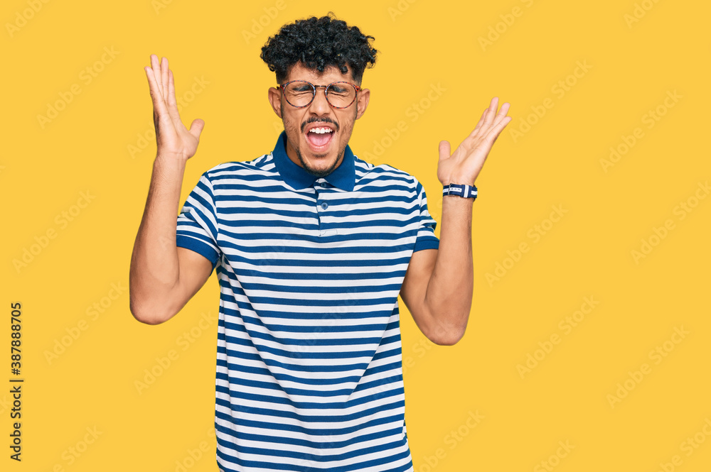 Young arab man wearing casual clothes and glasses celebrating mad and crazy for success with arms raised and closed eyes screaming excited. winner concept Young arab man wearing casual clothes and glasses celebrating mad and crazy for success with arms raised and closed eyes screaming excited. winner concept