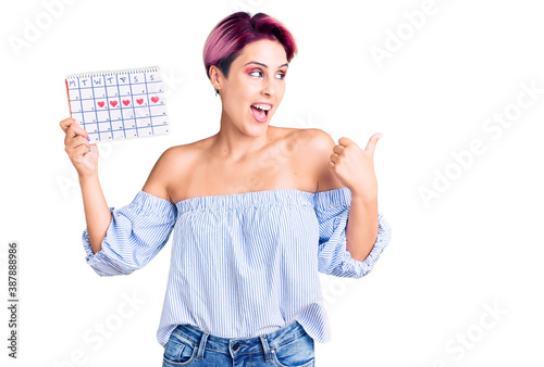 Young beautiful woman with pink hair holding heart calendar pointing thumb up to the side smiling happy with open mouth photo