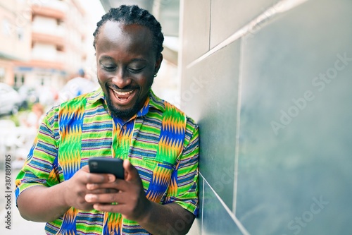 Young african american man using smartphone leaning on the wall at street of city.