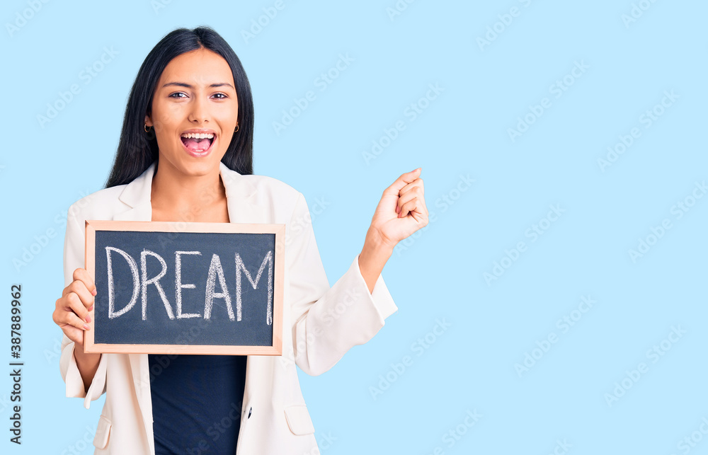 Young beautiful latin girl holding blackboard with dream word screaming proud, celebrating victory and success very excited with raised arms Young beautiful latin girl holding blackboard with dream word screaming proud, celebrating victory and success very excited with raised arms
