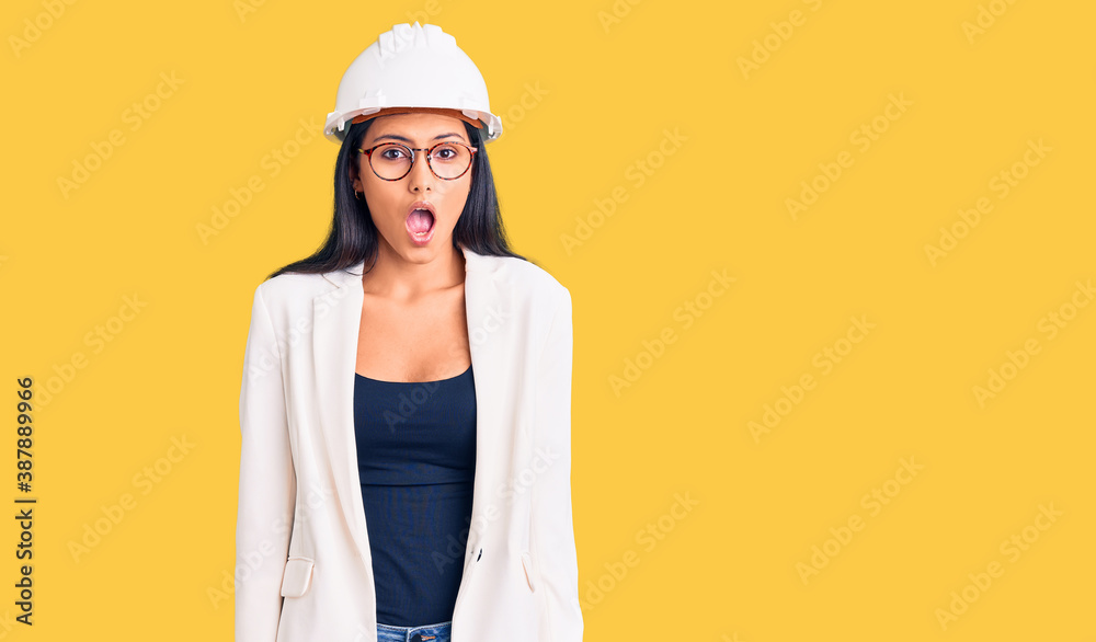 Young beautiful latin girl wearing architect hardhat and glasses afraid and shocked with surprise expression, fear and excited face. Young beautiful latin girl wearing architect hardhat and glasses afraid and shocked with surprise expression, fear and excited face.