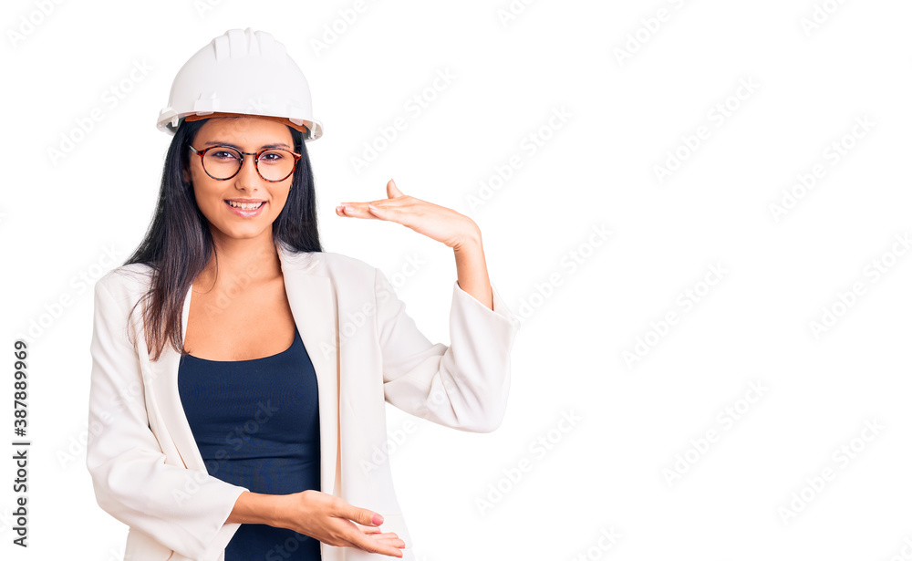 Young beautiful latin girl wearing architect hardhat and glasses gesturing with hands showing big and large size sign, measure symbol. smiling looking at the camera. measuring concept. Young beautiful latin girl wearing architect hardhat and glasses gesturing with hands showing big and large size sign, measure symbol. smiling looking at the camera. measuring concept.