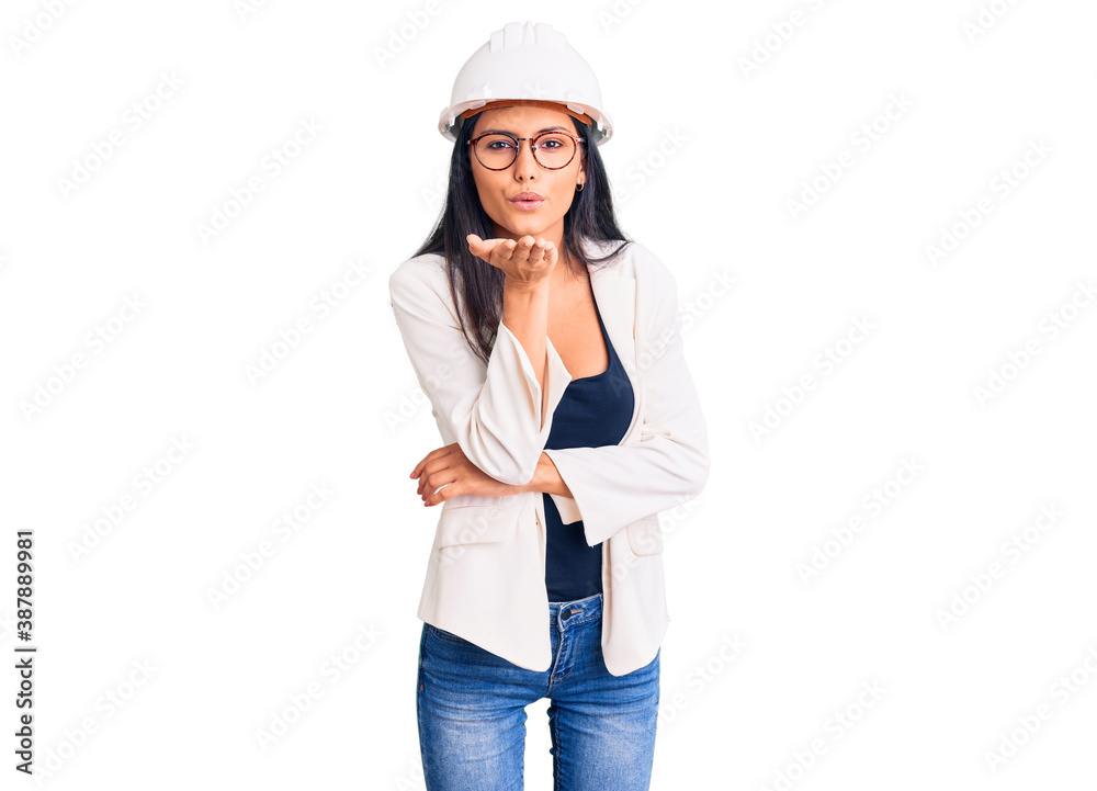 Young beautiful latin girl wearing architect hardhat and glasses looking at the camera blowing a kiss with hand on air being lovely and sexy. love expression.