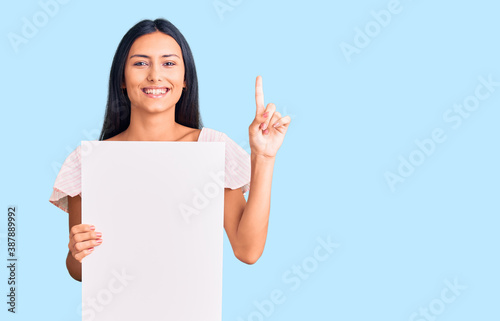 Young beautiful latin girl holding blank empty banner surprised with an idea or question pointing finger with happy face, number one
