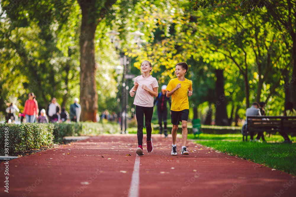 Fototapeta premium Couple of kids boy and girl doing cardio workout, jogging in park on jogging track red. Cute twins runing together. Run children, young athletes. Teen brother and sister running along path outdoors
