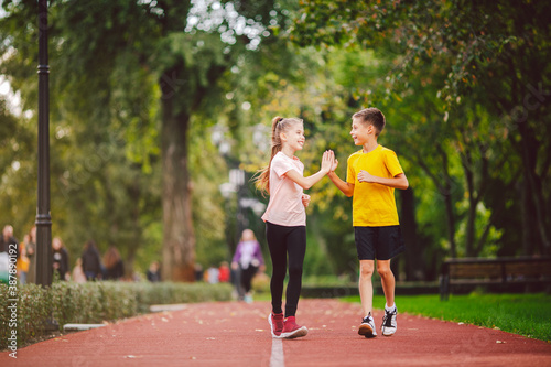 Couple of kids boy and girl doing cardio workout, jogging in park on jogging track red. Cute twins runing together. Run children, young athletes. Teen brother and sister running along path outdoors