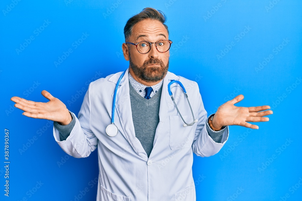 Handsome middle age man wearing doctor uniform and stethoscope clueless and confused expression with arms and hands raised. doubt concept.