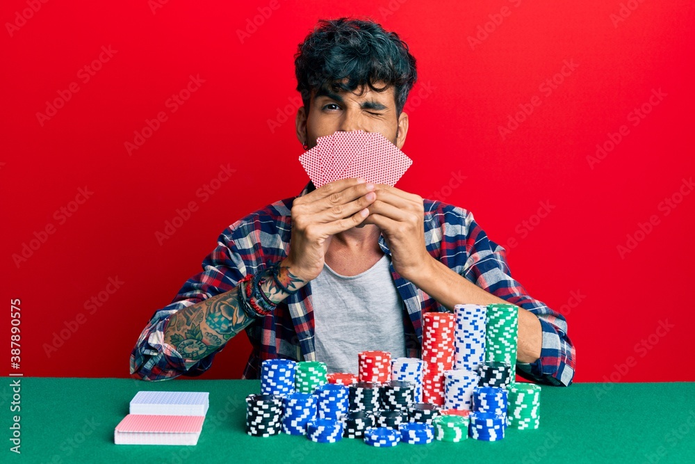 Young hispanic man playing gambling poker covering face with cards winking looking at the camera with sexy expression, cheerful and happy face. Young hispanic man playing gambling poker covering face with cards winking looking at the camera with sexy expression, cheerful and happy face.