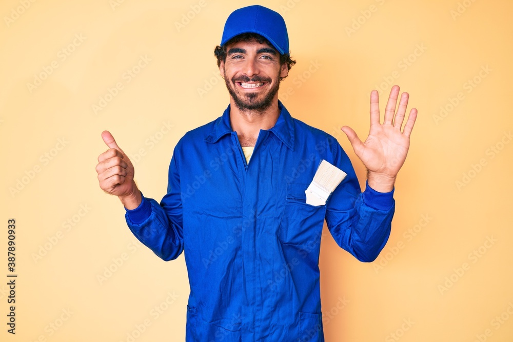 Handsome young man with curly hair and bear wearing builder jumpsuit uniform showing and pointing up with fingers number six while smiling confident and happy.