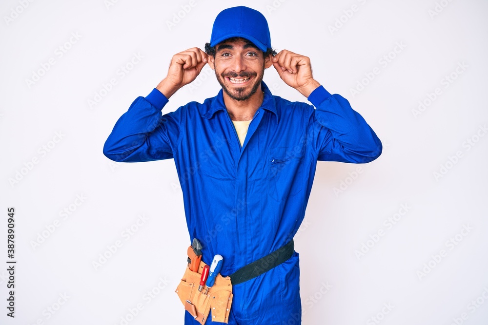 Handsome young man with curly hair and bear weaing handyman uniform smiling pulling ears with fingers, funny gesture. audition problem