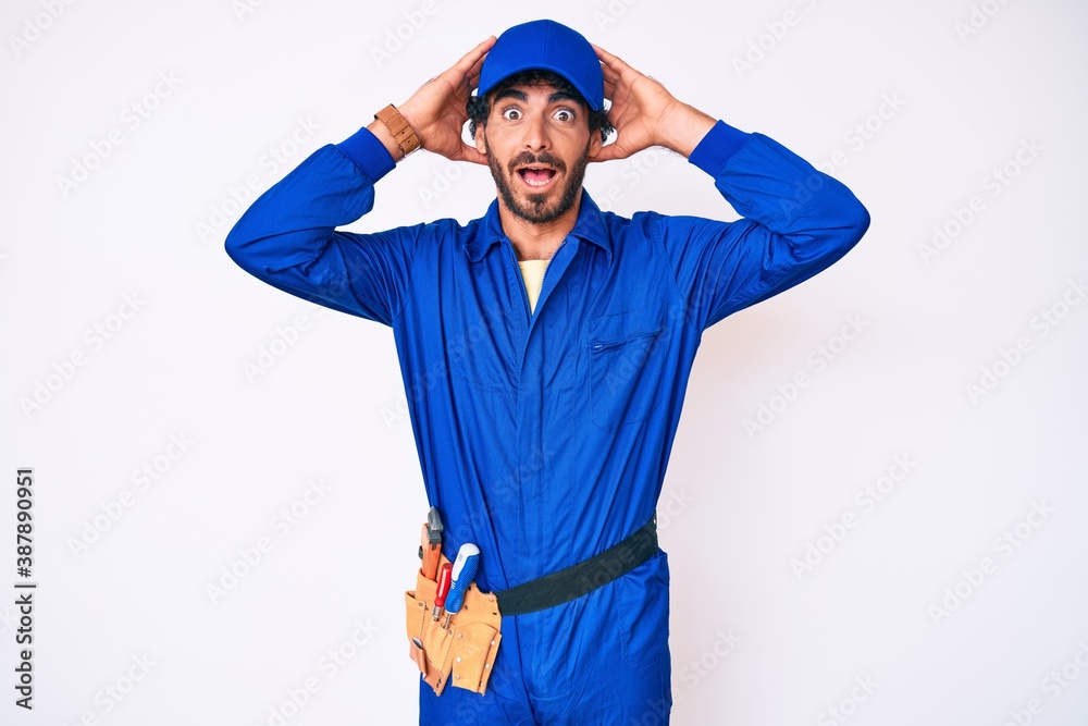 Handsome young man with curly hair and bear weaing handyman uniform crazy and scared with hands on head, afraid and surprised of shock with open mouth