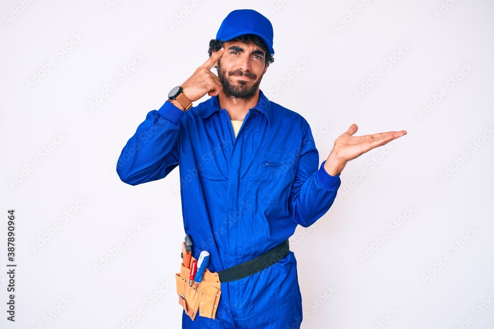 Handsome young man with curly hair and bear weaing handyman uniform confused and annoyed with open palm showing copy space and pointing finger to forehead. think about it.
