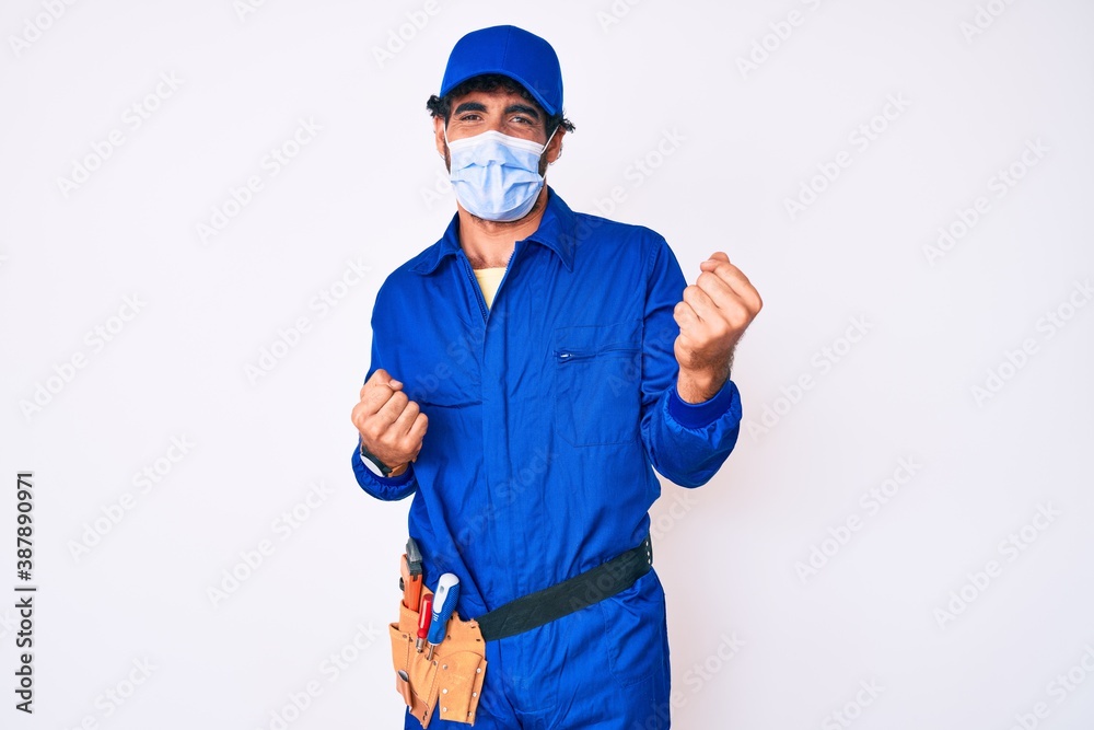 Handsome young man with curly hair and bear wearing handyman uniform and covid-19 safety mask celebrating surprised and amazed for success with arms raised and eyes closed